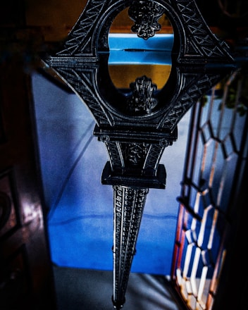 An intricately designed metal or wooden structure with geometric patterns and ornate detailing, possibly part of an architectural feature or decorative object. The background includes a stained glass window with colorful panes, casting a multi-hued light.