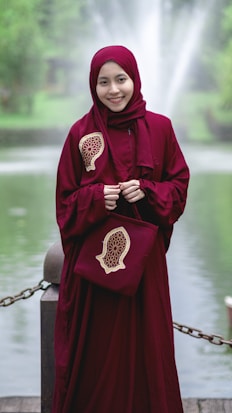 A person wearing a deep red hijab and dress stands cheerfully in front of a serene pond with a fountain in the background. The outfit features intricate designs on the shoulder and matching bag, giving an elegant touch. Greenery surrounds the area, contributing to a peaceful ambiance.