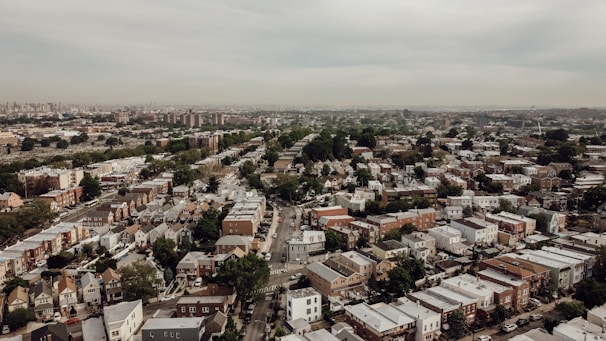 Aerial view of a sprawling suburban neighborhood with rows of houses and tree-lined streets extending to the horizon. The architecture features a mix of styles, predominantly low-rise residential buildings with white or light-colored roofs. The area is densely packed, with larger apartment buildings visible in the distance, and a muted sky overhead.