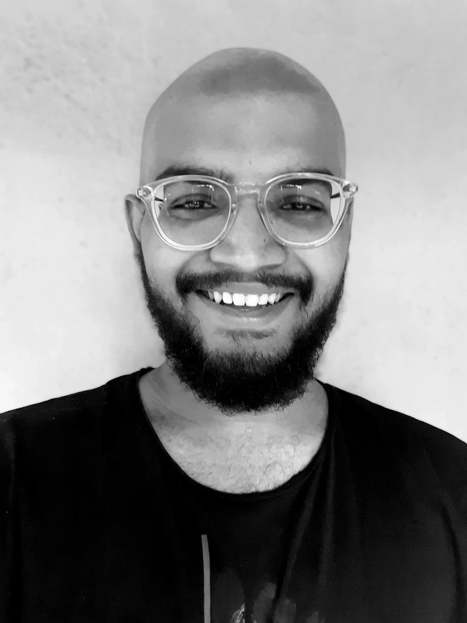 The image shows a man with a bald head and glasses, wearing a black t-shirt. He is smiling at the camera, conveying a sense of joy and positivity. The photograph is taken from a low angle, making him appear larger than life against the backdrop of a white wall.