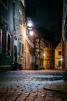 A cobblestone street in a narrow alley at night is dimly lit by warm street lights. The buildings on either side have a rustic charm, featuring wooden beams and flower boxes on the windows. A sign for a hotel hangs near one of the lamps, illuminating the area with a soft glow.