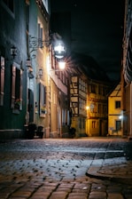 A cobblestone street in a narrow alley at night is dimly lit by warm street lights. The buildings on either side have a rustic charm, featuring wooden beams and flower boxes on the windows. A sign for a hotel hangs near one of the lamps, illuminating the area with a soft glow.