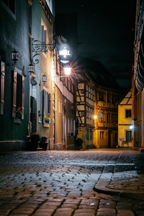 A cobblestone street in a narrow alley at night is dimly lit by warm street lights. The buildings on either side have a rustic charm, featuring wooden beams and flower boxes on the windows. A sign for a hotel hangs near one of the lamps, illuminating the area with a soft glow.