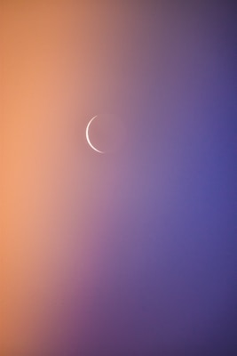 A thin crescent moon set against a gradient background transitioning from soft orange to deep blue. The scene gives a tranquil and serene appearance, emphasizing the delicate and subtle presence of the moon.