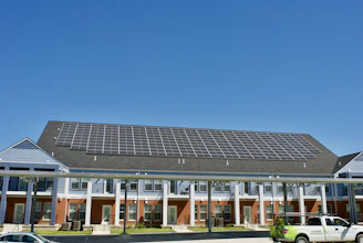 A building with a large array of solar panels on its roof. The structure is multi-story, featuring red brick on the lower level and blue siding on the upper parts. There are multiple entrances along the front, with a covered walkway supported by columns. A clear blue sky is visible in the background. A white truck is parked in front.