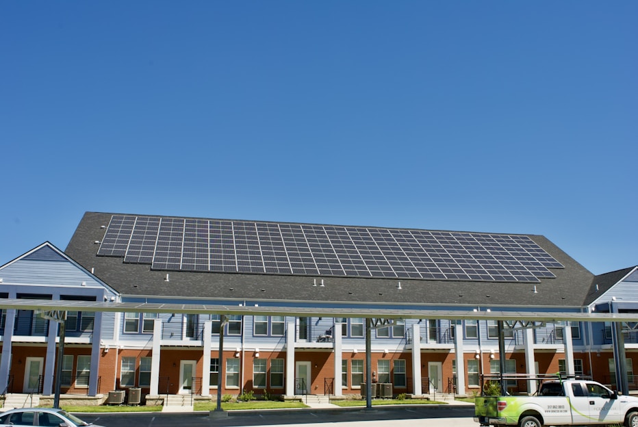 A building with a large array of solar panels on its roof. The structure is multi-story, featuring red brick on the lower level and blue siding on the upper parts. There are multiple entrances along the front, with a covered walkway supported by columns. A clear blue sky is visible in the background. A white truck is parked in front.