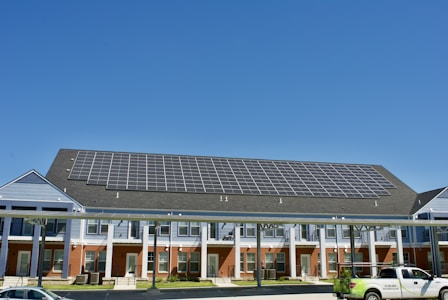A building with a large array of solar panels on its roof. The structure is multi-story, featuring red brick on the lower level and blue siding on the upper parts. There are multiple entrances along the front, with a covered walkway supported by columns. A clear blue sky is visible in the background. A white truck is parked in front.
