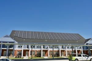A building with a large array of solar panels on its roof. The structure is multi-story, featuring red brick on the lower level and blue siding on the upper parts. There are multiple entrances along the front, with a covered walkway supported by columns. A clear blue sky is visible in the background. A white truck is parked in front.