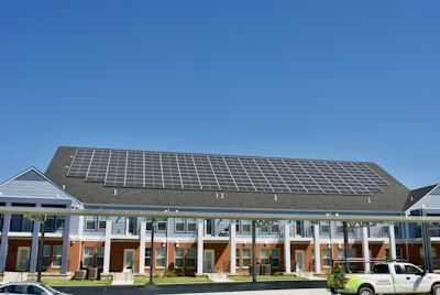 A building with a large array of solar panels on its roof. The structure is multi-story, featuring red brick on the lower level and blue siding on the upper parts. There are multiple entrances along the front, with a covered walkway supported by columns. A clear blue sky is visible in the background. A white truck is parked in front.