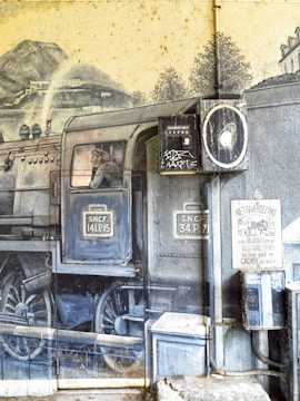 A realistic mural of a steam locomotive is painted on a wall, featuring intricate details such as wheels, smoke, and mechanical parts. A person is depicted inside the train, looking out from a window. There are several signs, utilities, and graffiti painted or attached on and around the mural, adding layers to the urban artwork.