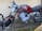 A vintage motorcycle with a red fuel tank and rust on various metal parts is parked against a textured beige wall. The motorcycle has a round headlight and is missing some parts, indicating its age and wear. The ground below is bare, with some grass and debris scattered.