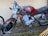 A vintage motorcycle with a red fuel tank and rust on various metal parts is parked against a textured beige wall. The motorcycle has a round headlight and is missing some parts, indicating its age and wear. The ground below is bare, with some grass and debris scattered.