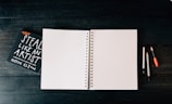 An open blank spiral notebook is placed on a dark wooden surface. To the left, there is a book titled 'Steal Like an Artist' by Austin Kleon. On the right side of the notebook, there are three writing instruments: two pencils and one pen, each with different colored caps.
