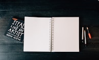 An open blank spiral notebook is placed on a dark wooden surface. To the left, there is a book titled 'Steal Like an Artist' by Austin Kleon. On the right side of the notebook, there are three writing instruments: two pencils and one pen, each with different colored caps.