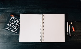 An open blank spiral notebook is placed on a dark wooden surface. To the left, there is a book titled 'Steal Like an Artist' by Austin Kleon. On the right side of the notebook, there are three writing instruments: two pencils and one pen, each with different colored caps.