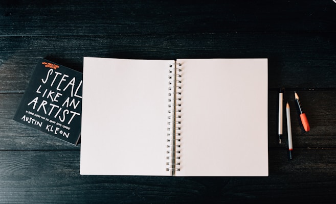 An open blank spiral notebook is placed on a dark wooden surface. To the left, there is a book titled 'Steal Like an Artist' by Austin Kleon. On the right side of the notebook, there are three writing instruments: two pencils and one pen, each with different colored caps.