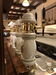 Several ornate beer taps with white ceramic and gold accents are lined up on a bar counter. Above the bar, hanging lights with warm shades illuminate the area. Shelves in the background display an assortment of bottles, and glassware is neatly arranged underneath. The wooden beams on the ceiling add a rustic touch to the setting.