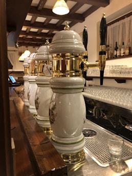 Several ornate beer taps with white ceramic and gold accents are lined up on a bar counter. Above the bar, hanging lights with warm shades illuminate the area. Shelves in the background display an assortment of bottles, and glassware is neatly arranged underneath. The wooden beams on the ceiling add a rustic touch to the setting.