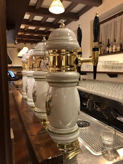 Several ornate beer taps with white ceramic and gold accents are lined up on a bar counter. Above the bar, hanging lights with warm shades illuminate the area. Shelves in the background display an assortment of bottles, and glassware is neatly arranged underneath. The wooden beams on the ceiling add a rustic touch to the setting.