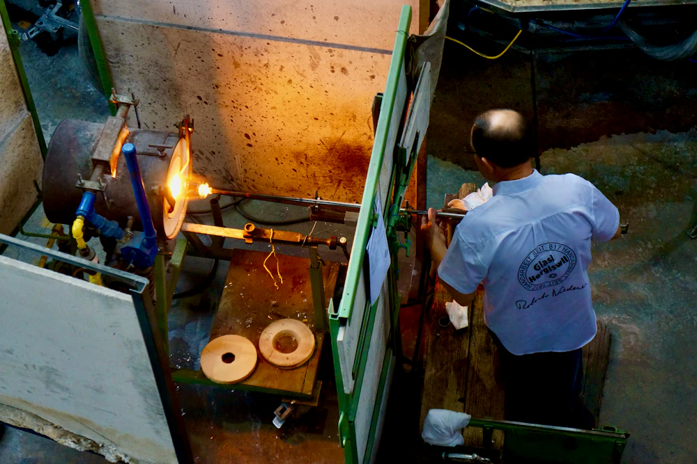 Two people working together at a glass furnace station, one guiding the other