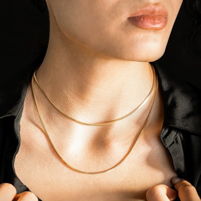 A close-up of a person's neck adorned with two thin gold necklaces, wearing a black shirt. The image focuses on the elegant simplicity of the jewelry against the skin, highlighting texture and light.
