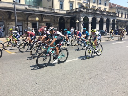 A group of cyclists in colorful uniforms is participating in a road race on a city street. The background features a historic building with arches and several onlookers watching from the sidewalk.