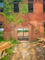 A partially demolished brick building with exposed interior and remnants of wooden debris. Green ivy covers parts of the wall, and there is an open doorway leading to a pile of rubble. The ground is uneven with scattered construction materials.