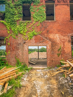 A partially demolished brick building with exposed interior and remnants of wooden debris. Green ivy covers parts of the wall, and there is an open doorway leading to a pile of rubble. The ground is uneven with scattered construction materials.