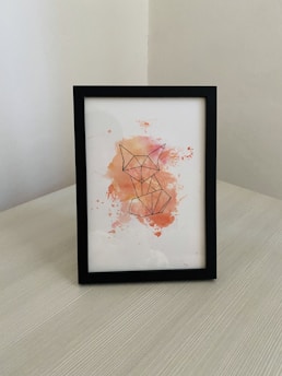 A framed piece of artwork featuring a geometric outline of a fox. The illustration is rendered on a white background with watercolor splashes in shades of orange, pink, and yellow surrounding the fox outline. The frame is black and placed on a light-colored wooden surface, against a corner background with white walls.