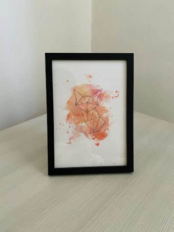 A framed piece of artwork featuring a geometric outline of a fox. The illustration is rendered on a white background with watercolor splashes in shades of orange, pink, and yellow surrounding the fox outline. The frame is black and placed on a light-colored wooden surface, against a corner background with white walls.