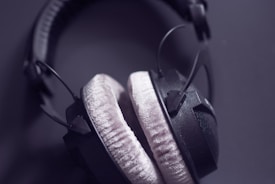 A pair of over-ear headphones with soft, plush ear cushions. The headphones are predominantly black with padded headband and visible wires on a dark background.