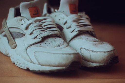 Worn white sneakers with visible dirt and creases are placed on a wooden surface. The laces are loosely tied and the shoes have a retro style.