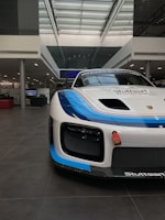 A sleek, white race car with a blue and orange stripe detail is positioned inside an industrial showroom with a high glass ceiling. The car has a prominent front bumper and a sporty design, with brand logos visible.