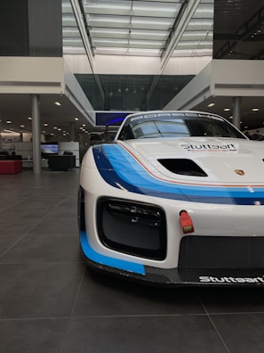 A sleek, white race car with a blue and orange stripe detail is positioned inside an industrial showroom with a high glass ceiling. The car has a prominent front bumper and a sporty design, with brand logos visible.
