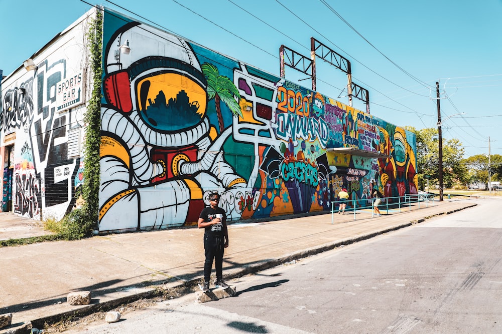 Mann in schwarzer Jacke und schwarzer Hose geht tagsüber auf dem Bürgersteig in der Nähe der Graffiti-Wand spazieren