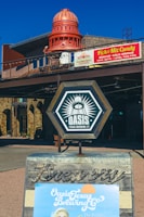 A rustic outdoor setting featuring a prominent brewery sign with the logo for 'Oasis Texas Brewing Co' displayed on a wooden A-frame. In the background, there is a building with a large decorative red dome structure. A sign nearby advertises a 'Pick & Mix Candy' with various snacks and gifts.