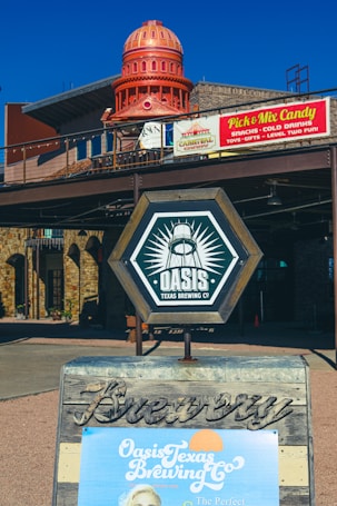 A rustic outdoor setting featuring a prominent brewery sign with the logo for 'Oasis Texas Brewing Co' displayed on a wooden A-frame. In the background, there is a building with a large decorative red dome structure. A sign nearby advertises a 'Pick & Mix Candy' with various snacks and gifts.