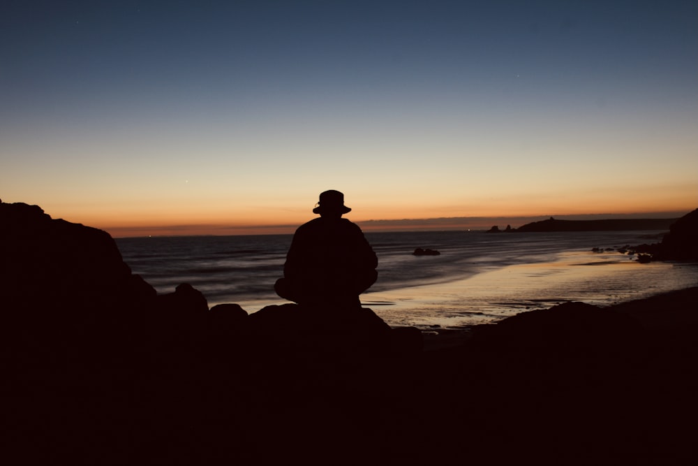 silhouette of man sitting on rock near sea during sunset