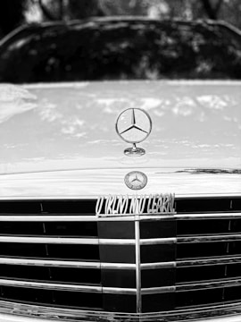 A close-up of a car hood shows a shiny emblem prominently featured on top, with a distinct star-shaped logo. The grille has horizontal slats and there is a badge with a circular design. The image is in black and white, giving a classic and sleek appearance.