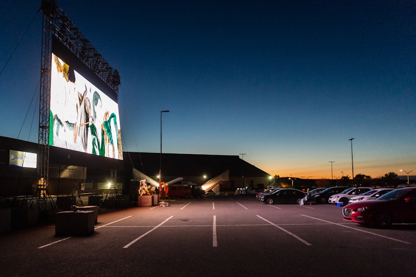 A large outdoor movie screen is illuminated showing a colorful image during twilight. In front of the screen, several cars are parked in an orderly fashion in a parking lot under a darkening sky with a hint of orange at the horizon, indicating the setting sun.