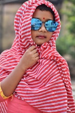 A child is wrapped in a red and white striped fabric, wearing reflective blue sunglasses. The fabric covers the hair and frames the face, with a small red dot on the forehead. The background is blurred, creating a focus on the child.