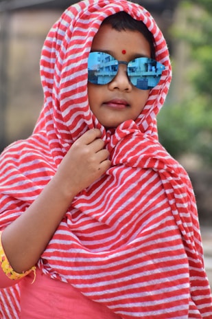 A child is wrapped in a red and white striped fabric, wearing reflective blue sunglasses. The fabric covers the hair and frames the face, with a small red dot on the forehead. The background is blurred, creating a focus on the child.