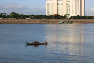 A small wooden boat is floating on a calm river, with two people on board. In the background, a large white high-rise building is visible, reflecting in the water. There is also a row of trees along the riverbank, and a larger green boat further away.