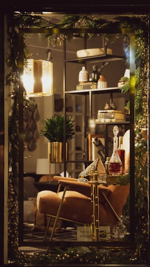 The image captures a cozy and elegantly decorated interior space, showcasing a warm lighting fixture, a plush armchair, and a tastefully arranged bookshelf adorned with decorative items. A decanter with a deep amber liquid sits on a stylish side table, accompanied by a patterned cushion and a healthy green plant in a gold pot. Festive greenery drapes over the frame, enhancing the ambiance.
