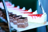 Several slices of dessert are arranged on a tiered stand. The desserts include pieces of chocolate cake with a glossy layer of frosting on top and slices of cheesecake topped with strawberry sauce.