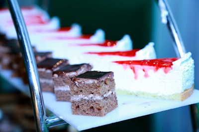 Several slices of dessert are arranged on a tiered stand. The desserts include pieces of chocolate cake with a glossy layer of frosting on top and slices of cheesecake topped with strawberry sauce.
