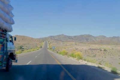 A long stretch of road stretches into the distance, surrounded by dry, rugged terrain with mountains in the background. A truck loaded with goods is visible on the left side of the image, partially in view, driving along the road. Sparse vegetation is scattered along the roadside.