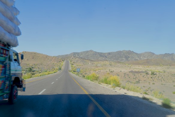 A long stretch of road stretches into the distance, surrounded by dry, rugged terrain with mountains in the background. A truck loaded with goods is visible on the left side of the image, partially in view, driving along the road. Sparse vegetation is scattered along the roadside.