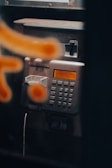 A close-up view of a metallic public payphone, featuring an alphanumeric keypad and an orange display screen. Some parts are out of focus due to a blurred orange pattern in the foreground.