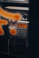 A close-up view of a metallic public payphone, featuring an alphanumeric keypad and an orange display screen. Some parts are out of focus due to a blurred orange pattern in the foreground.